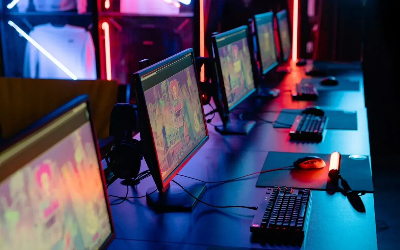 Rangée de postes de jeu dans une salle sombre éclairée au néon rouge et bleu, écrans allumés, claviers et casques posés sur les tables